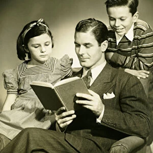Father reading to son (4-5) and daughter ( 6-7) in studio, (B&W), portrait